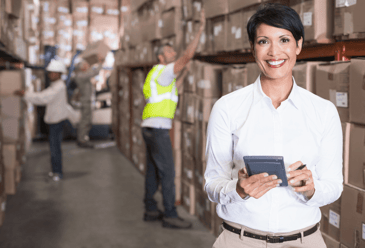 A cheerful warehouse manager using a tablet to effectively keep track of the numerous warehouse scanners and devices as her diligent team works in the background.